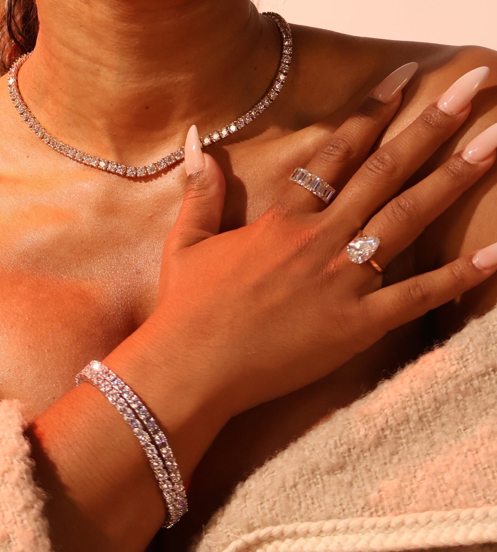 Close-up of a person wearing a diamond necklace, ring, and bracelet on a neutral background